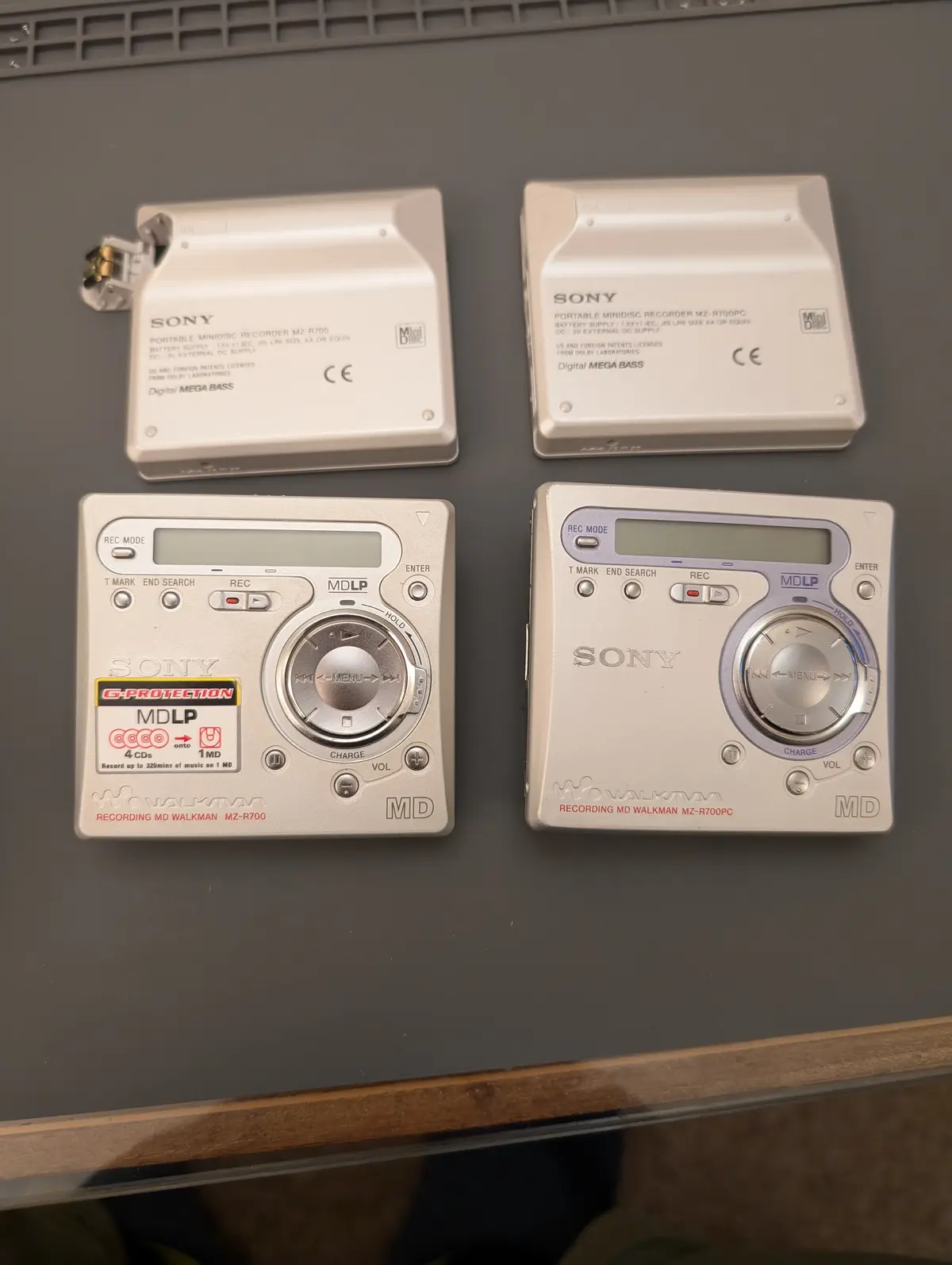 Four silver Sony Walkman MD recorders are arranged on a dark surface, two showing their backs with model numbers and…