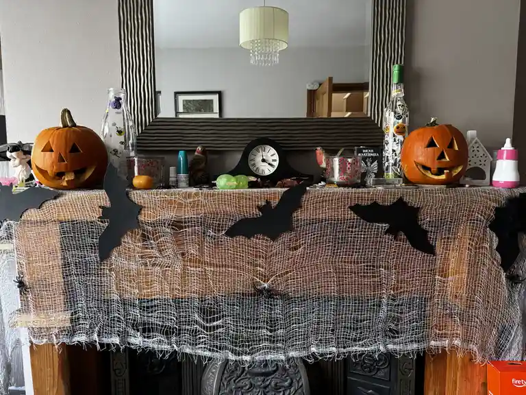 A mantelpiece is heavily decorated for Halloween with two carved pumpkins, black paper bats, white spiderweb fabric, and…