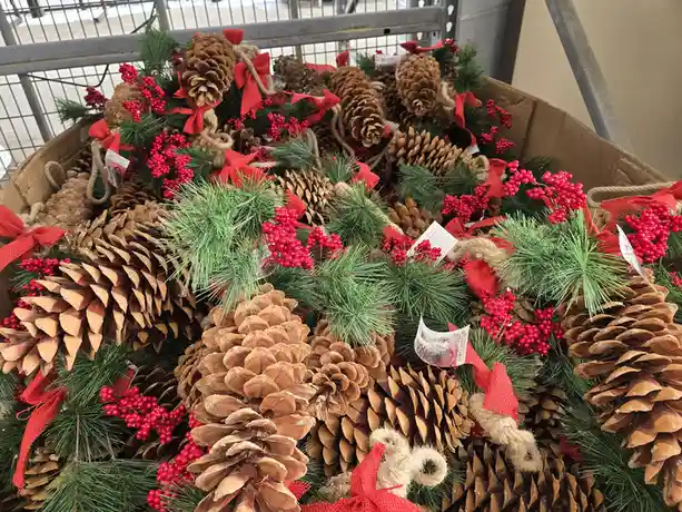 A large cardboard box is overflowing with holiday decorations, including numerous brown pinecones, green pine branches, and…