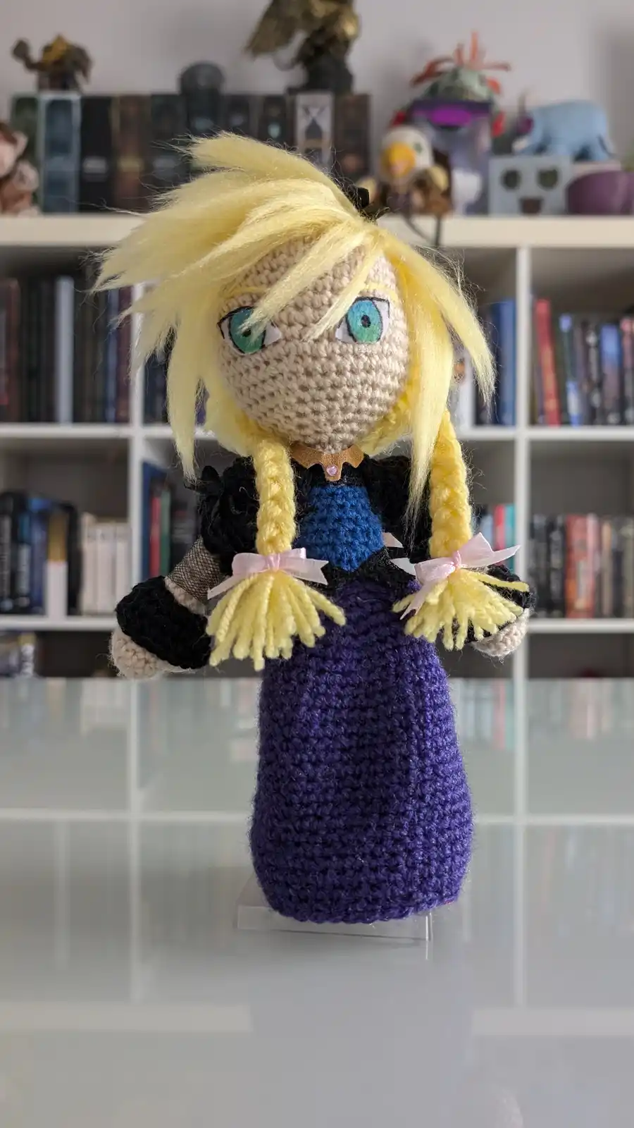 A handmade crochet doll with blonde hair in pigtails, blue eyes, and a purple dress with black sleeves stands on a clear…