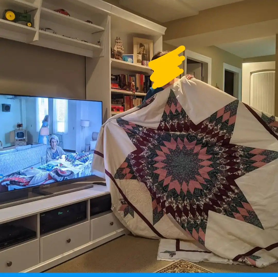 A person stands in a living room holding up a large, star-patterned quilt, partially obscuring their face with a yellow…