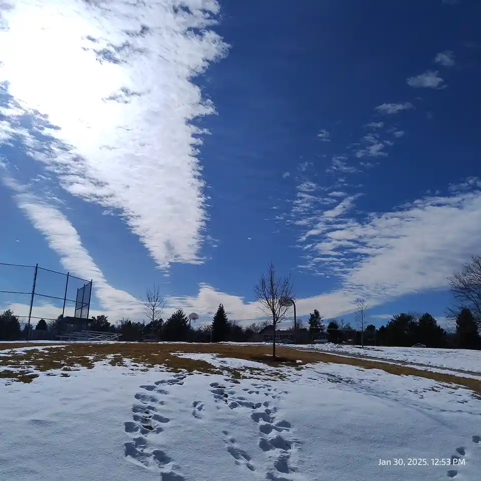 A snowy park scene with footprints leading across the snow toward a basketball hoop and chain-link fence under a bright blue…