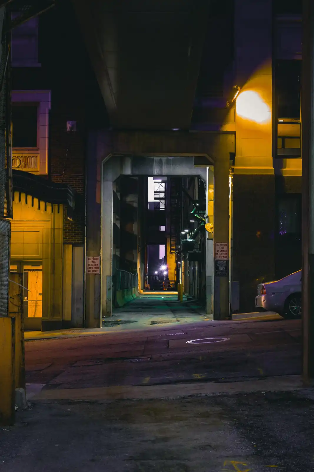 A nighttime urban alleyway under an overpass, lit by a warm yellow streetlamp, with a silver car parked on the right and a…