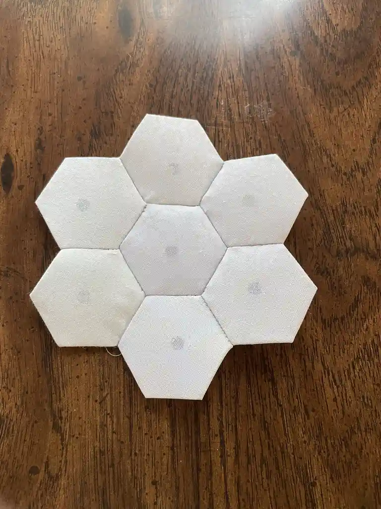 A handmade, light-colored fabric object composed of seven hexagonal pieces arranged in a flower-like pattern sits on a dark…
