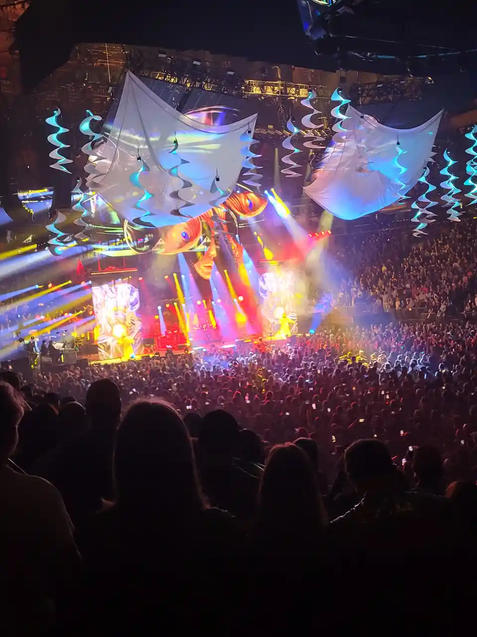 A wide-angle photo from inside a packed concert venue shows a massive audience facing a brightly lit stage with elaborate…