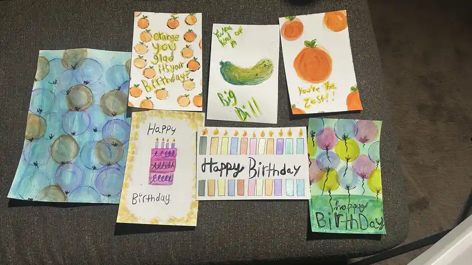 Seven handmade birthday cards lie on a gray fabric surface. Some feature watercolor balloons, a birthday cake, or candles…