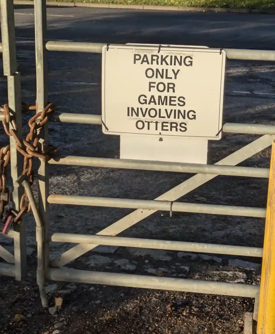 A white rectangular sign with black text reading 'PARKING ONLY FOR GAMES INVOLVING OTTERS' is taped to a metal gate. The…