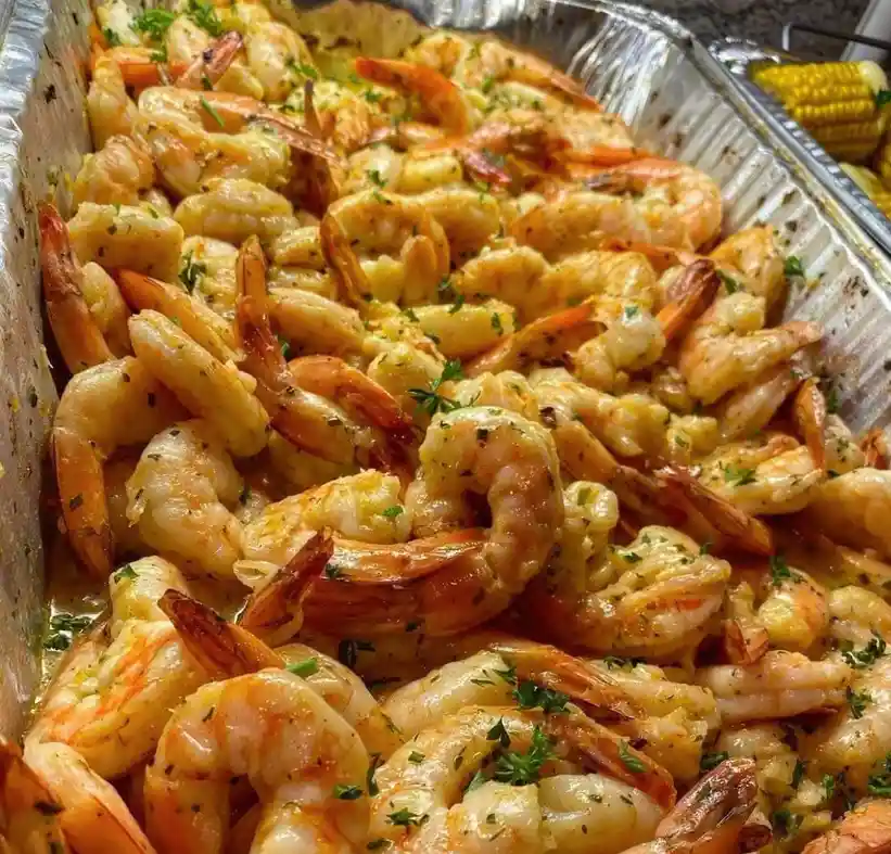 A close-up photo of a large aluminum tray filled with cooked, seasoned shrimp covered in a creamy sauce and garnished with…