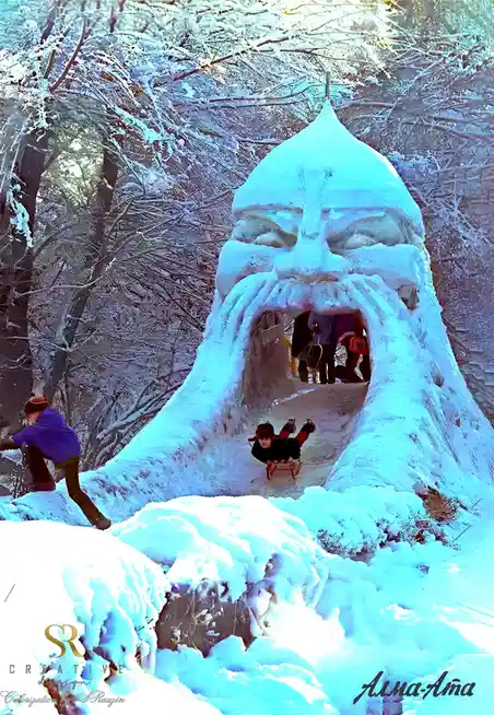 A large, stylized snow sculpture shaped like a giant's face with a wide mouth serving as a slide, set in a snowy forest. A…