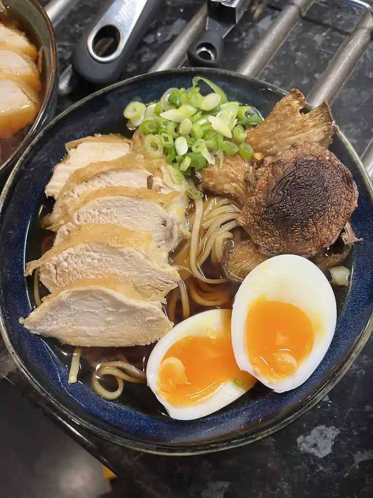 A close-up photo of a bowl of ramen with sliced chicken, mushrooms, a soft-boiled egg cut in half, and chopped green onions…