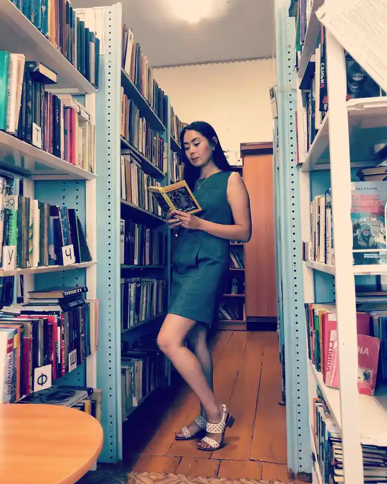 A woman in a green dress stands between tall bookshelves in a library, holding and reading a book. The shelves are packed…