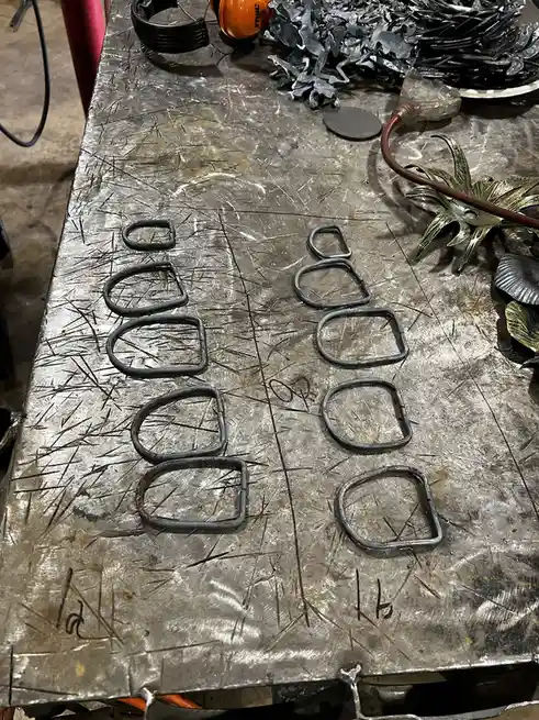 A metal workbench covered in scratches has two columns of handmade metal rings arranged like a comparison chart, labeled…
