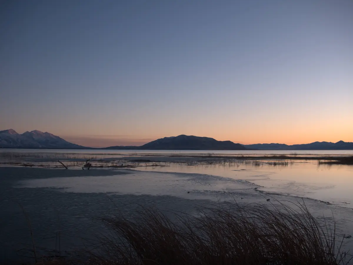 A serene landscape photo of a partially frozen lake at dusk, with snow-capped mountains in the distance under a gradient…