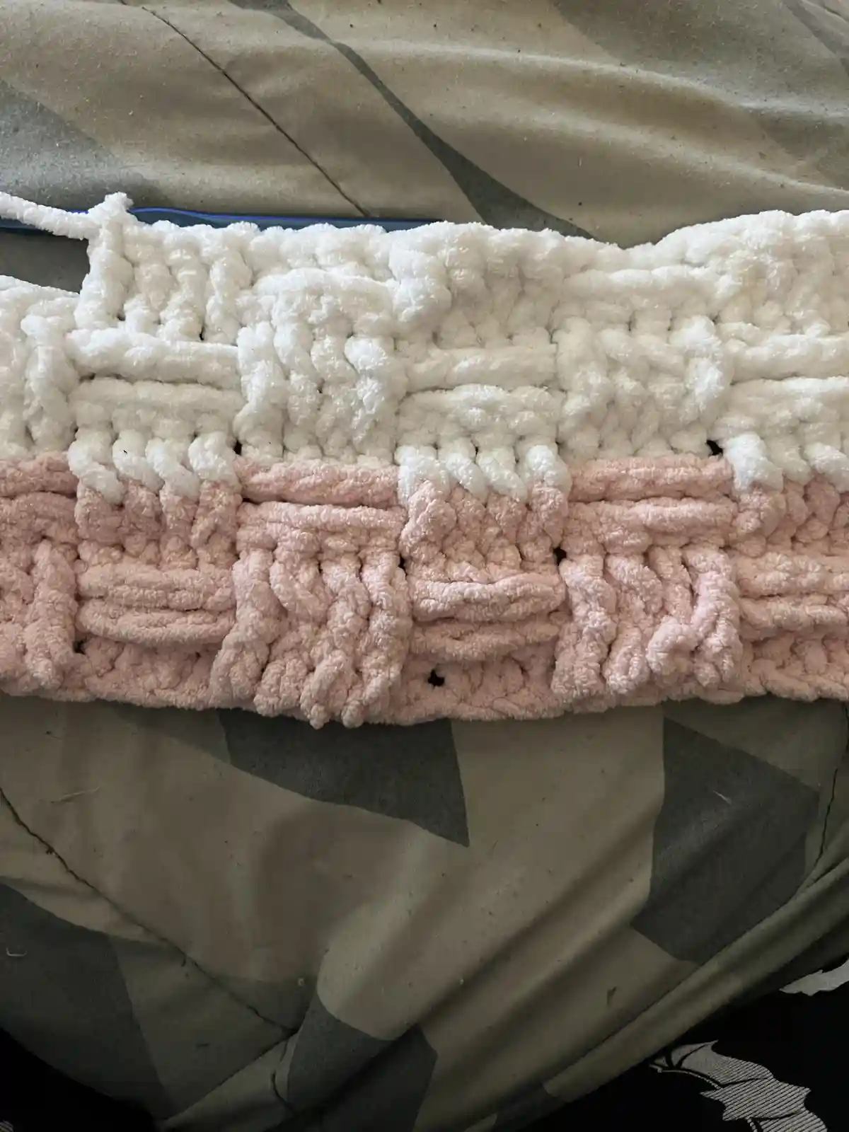 A close-up photo shows a chunky, textured crocheted blanket with a white top half and a pink bottom half, resting on a…
