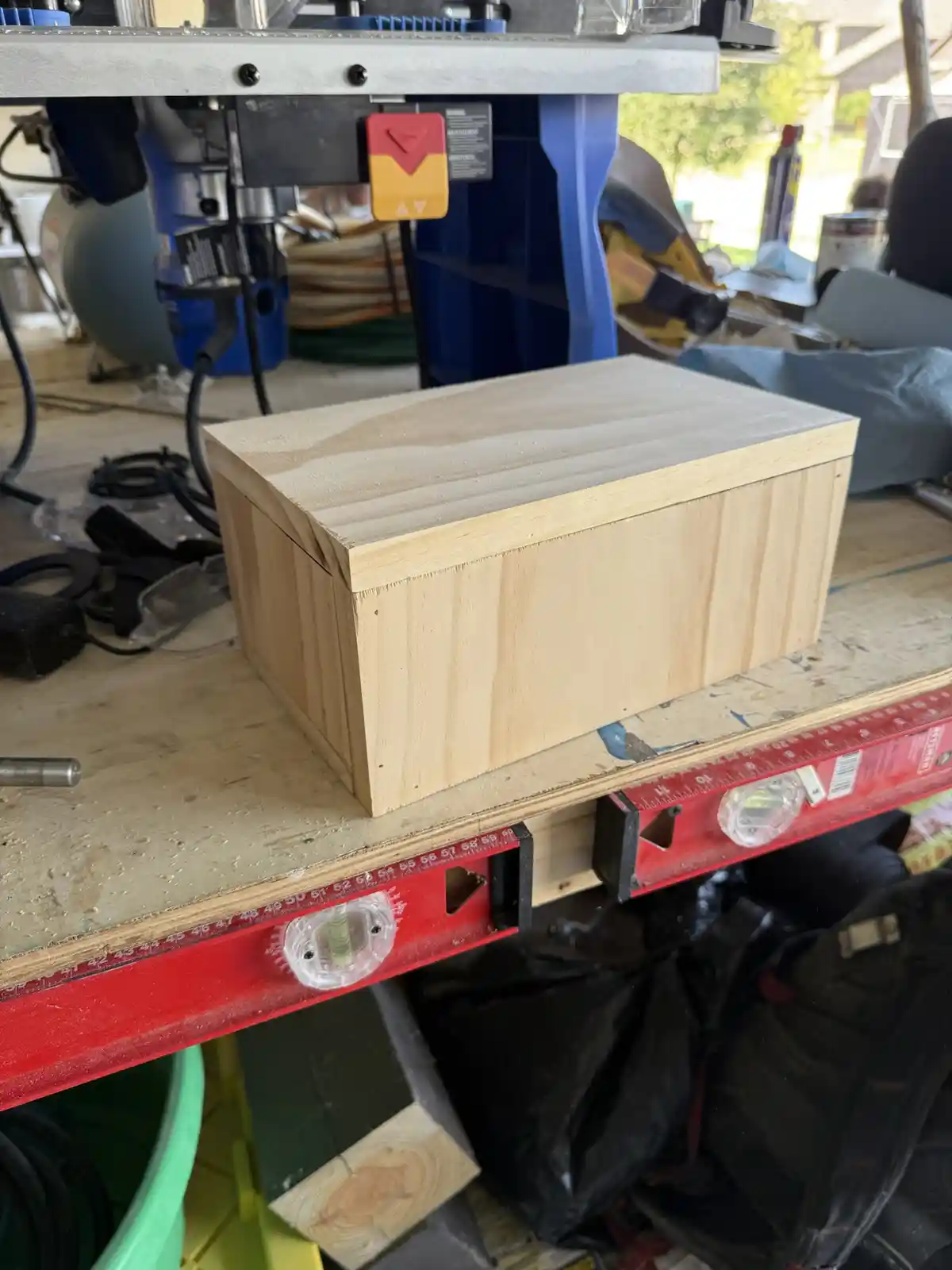 A light-colored wooden box sits on a workbench in a cluttered workshop, surrounded by tools and a red level. The box appears…