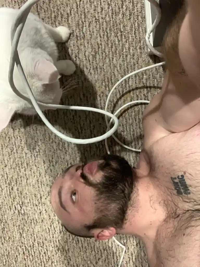 A shirtless man with a beard lies on a carpeted floor, looking up at a white cat whose paws are near tangled white cables…