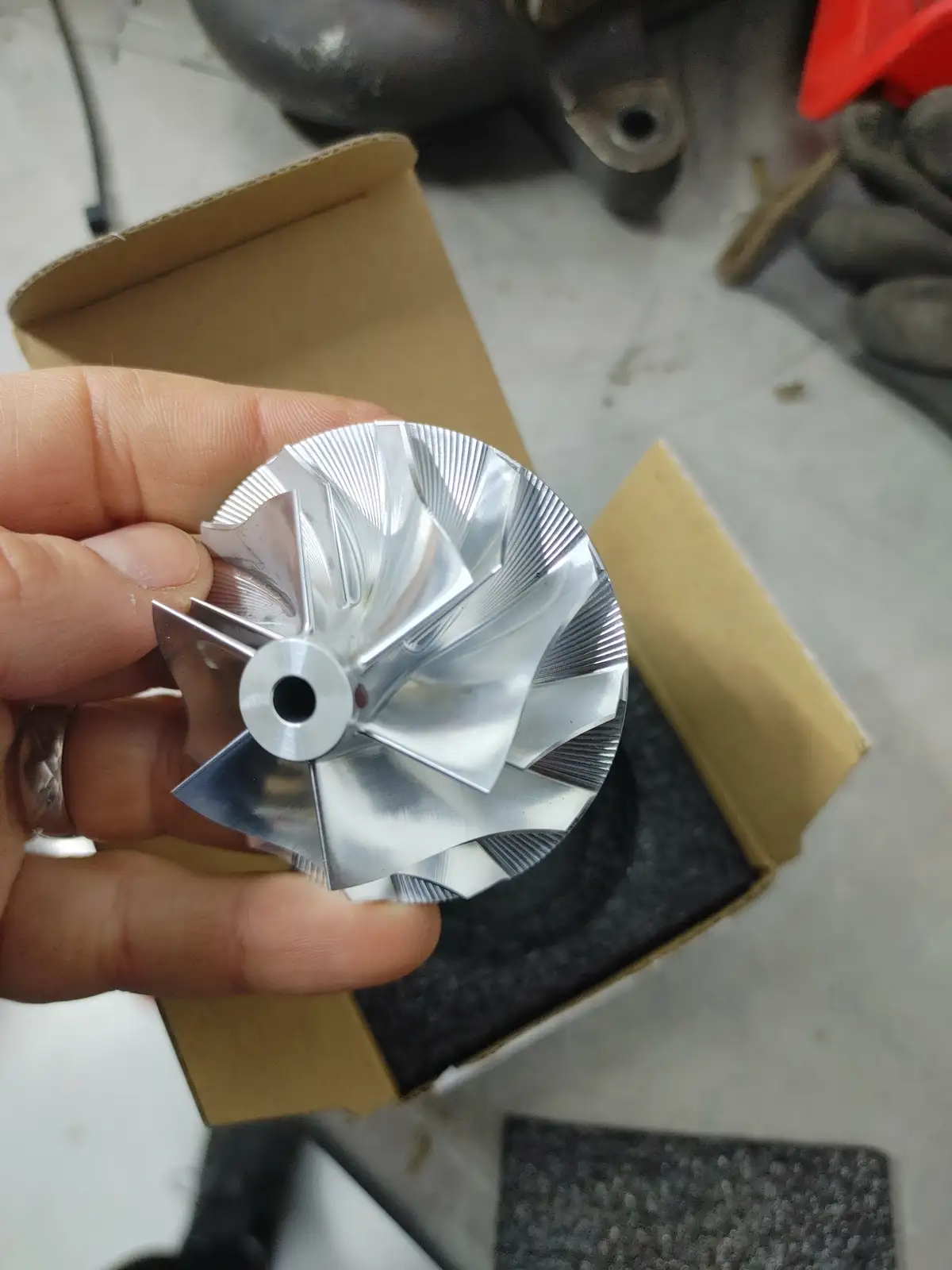 A hand holds a shiny, silver, turbine-like metal part with curved blades and a central hole, emerging from an open cardboard…