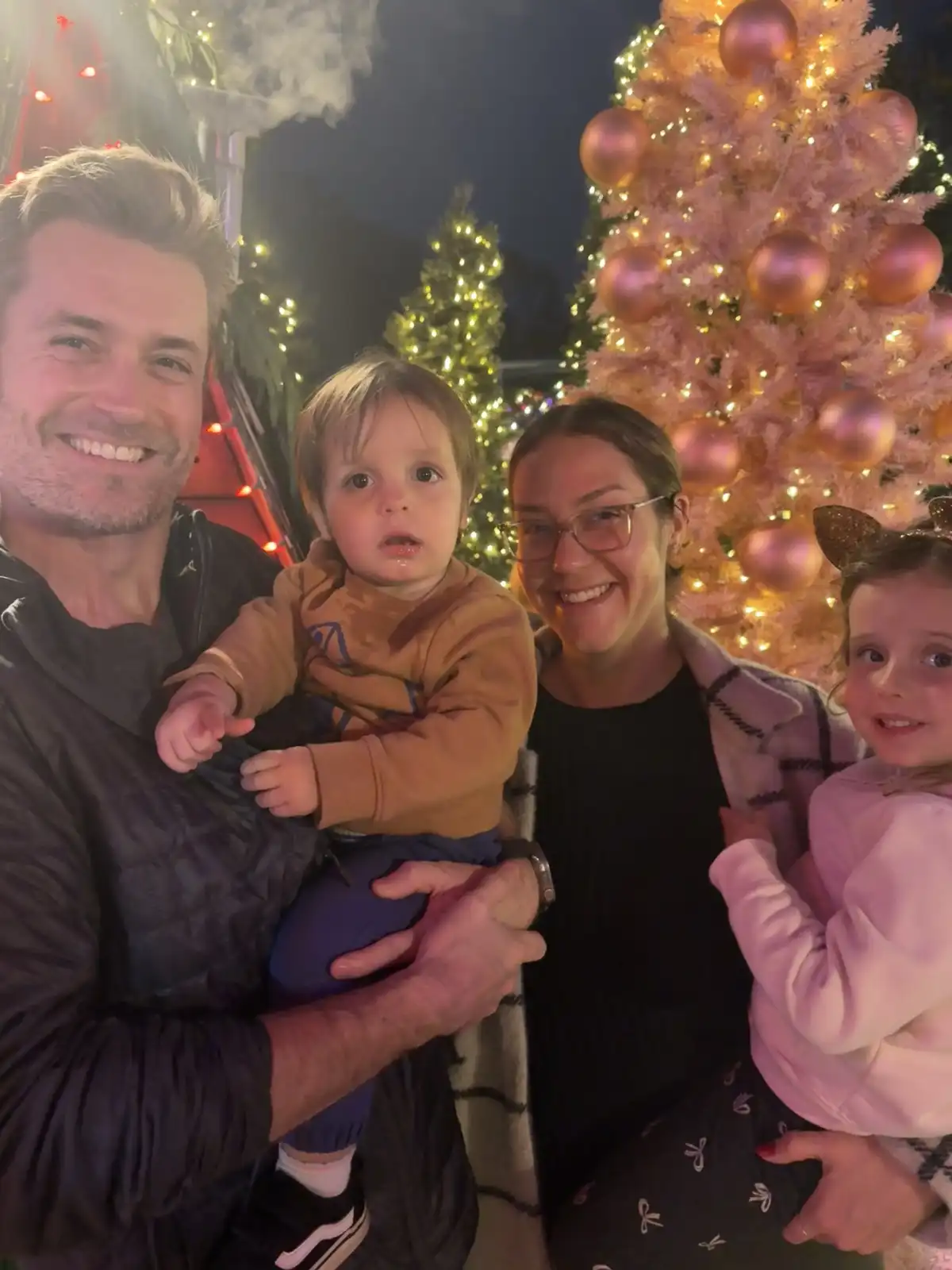 A family of four poses for a selfie at night in front of decorated Christmas trees, with the father holding a toddler and…
