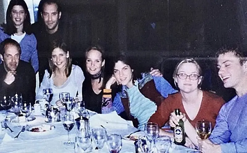 A grainy, low-light group photo of eight people at a dinner table with wine glasses and bottles, smiling or laughing. The…