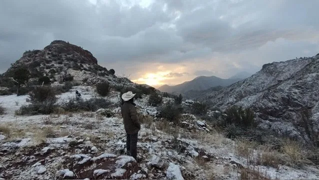 A person wearing a white hat and dark jacket stands on a snowy, rocky hillside looking out at a mountainous landscape under…