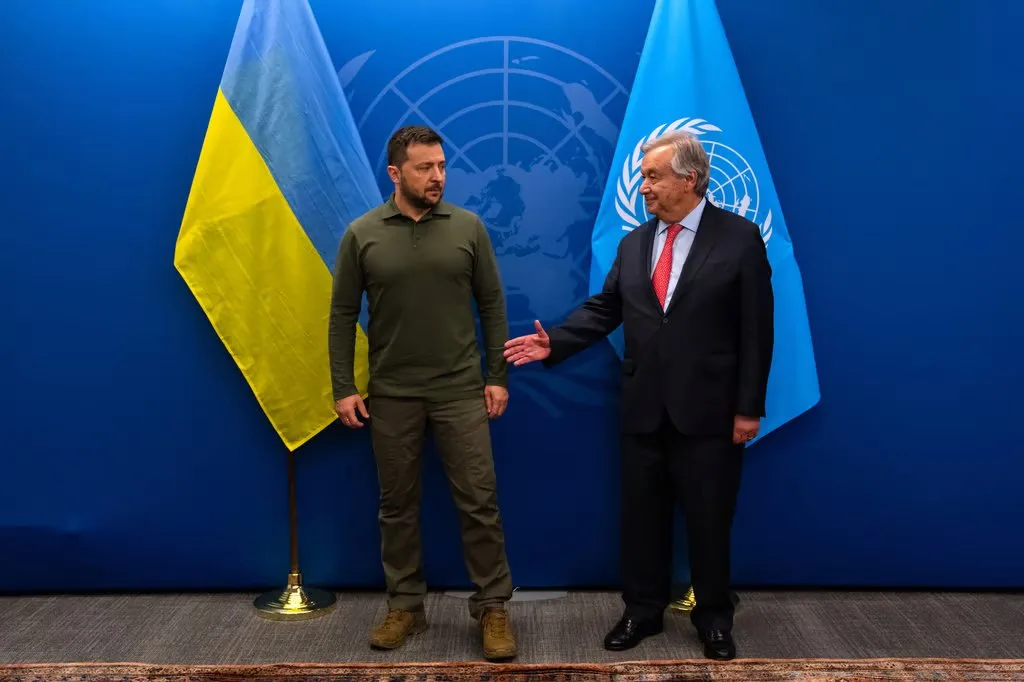 Two men stand facing each other in front of a blue backdrop with a UN logo and two flags: Ukraine on the left and the UN on…