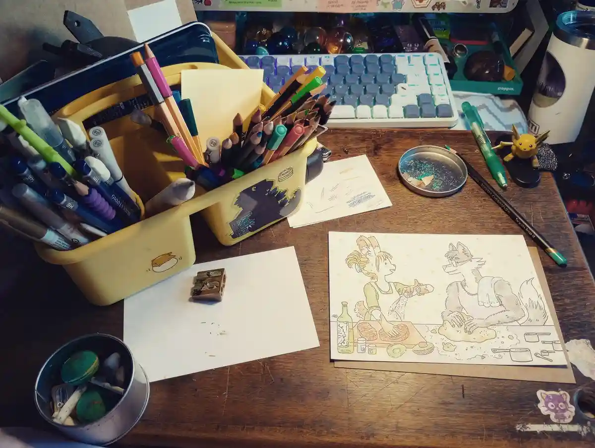 A cluttered artist's desk with a yellow pencil cup, a keyboard, and a drawing of two anthropomorphic characters cooking. The…