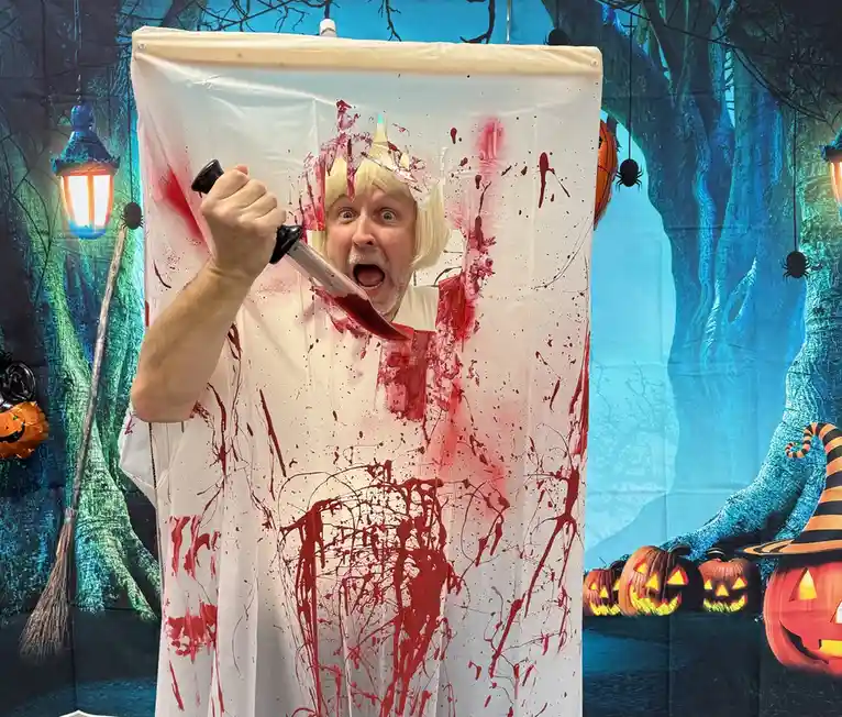 A man with a blonde wig and a surprised expression is holding a knife covered in red paint, emerging from a white curtain…