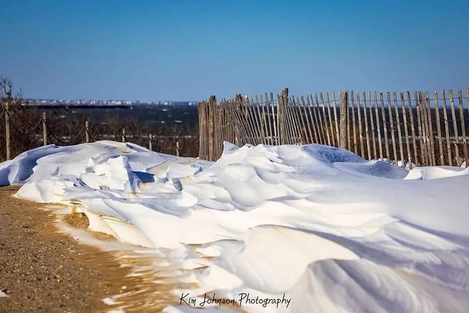 A large, uneven pile of melting snow lies on a dirt path next to a wooden fence under a clear blue sky. The snow is…