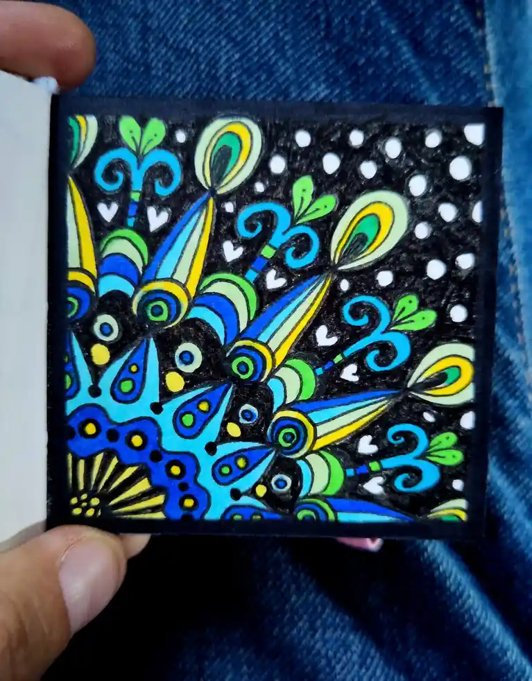 A hand holds a small, colorful, abstract drawing on black paper, featuring swirling patterns, teardrop shapes, and white…