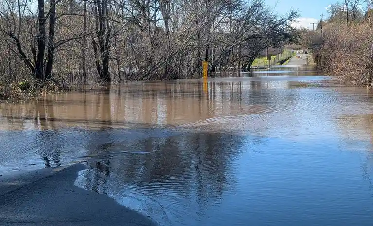 A flooded street with murky water covering the road, leaving only a small portion of the asphalt visible in the foreground…