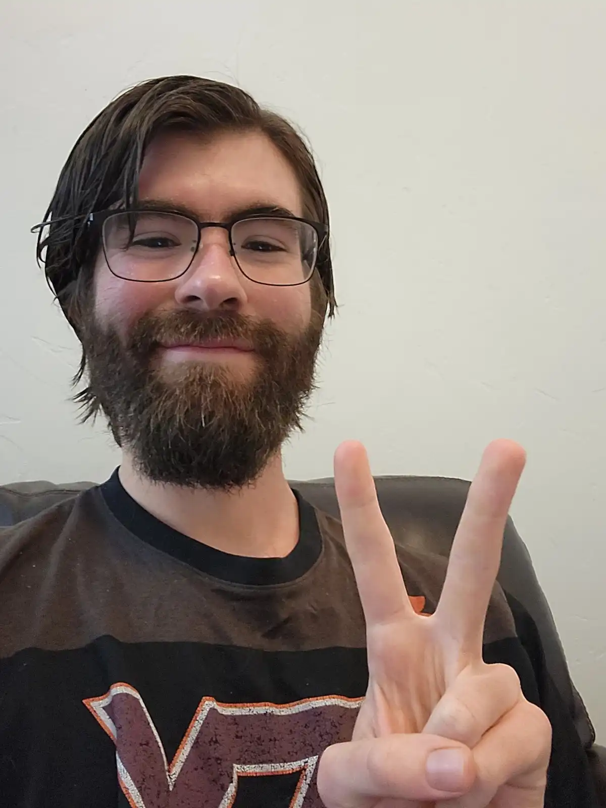 A man with a beard and glasses smiles while holding up a peace sign with his right hand. He wears a dark t-shirt with a…