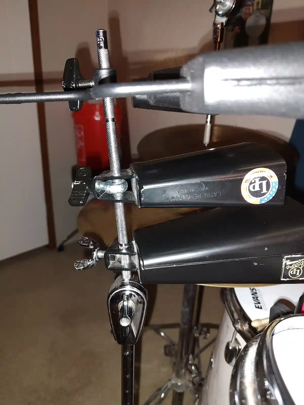 A close-up photo of a drum set's hardware, showing two black Latin Percussion cowbells mounted on a chrome stand with…