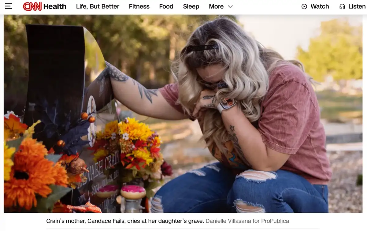 A woman with blonde hair and tattoos is kneeling at a grave marker, visibly crying with her head bowed. The grave is…