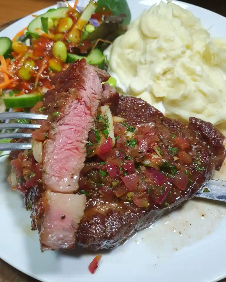 A close-up photo of a plate with a medium-rare steak being cut with a fork and knife, topped with a red onion and herb…