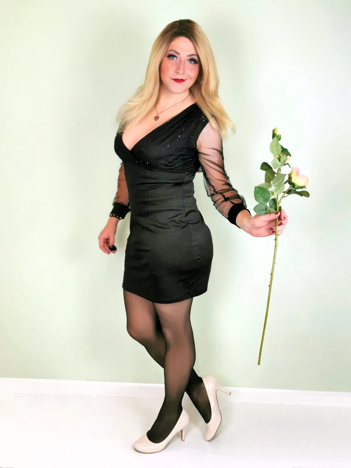 A woman with blonde hair poses in a black one-shoulder dress with sheer sleeves, black tights, and white heels, holding a…
