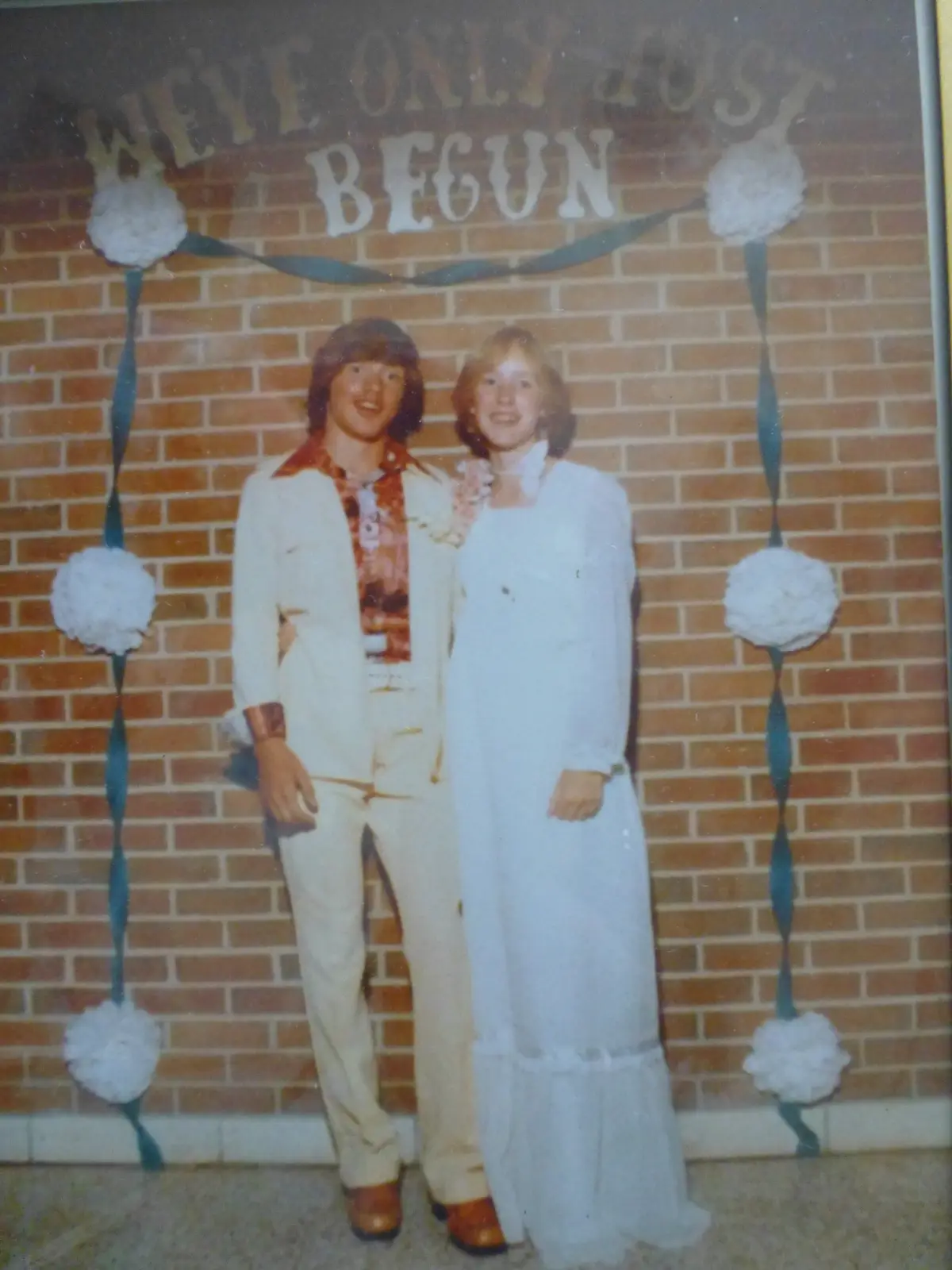 A vintage 1970s photo shows a young couple posing in front of a brick wall decorated with white paper pom-poms and blue…