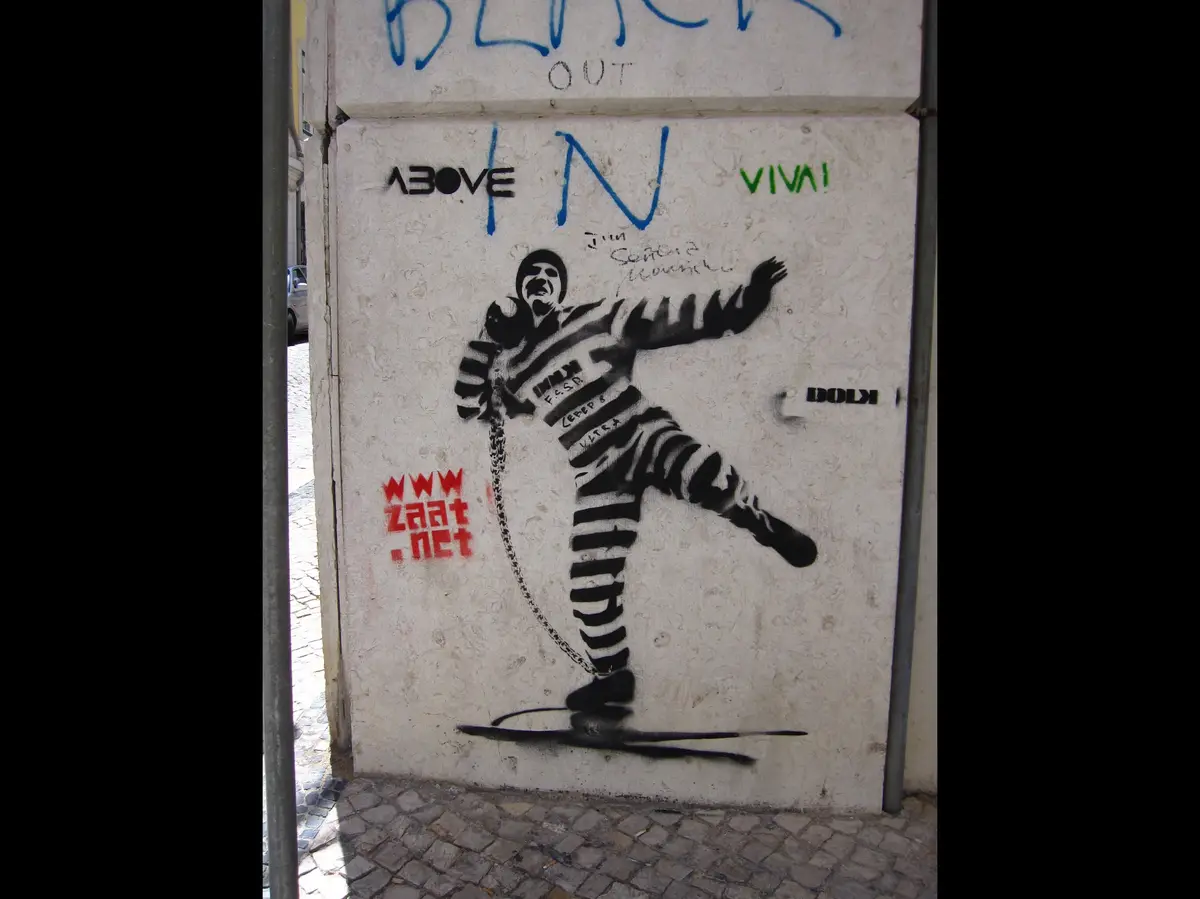 A black-and-white graffiti mural on a concrete wall depicts a prisoner in striped uniform, mid-kick, with a chain attached…