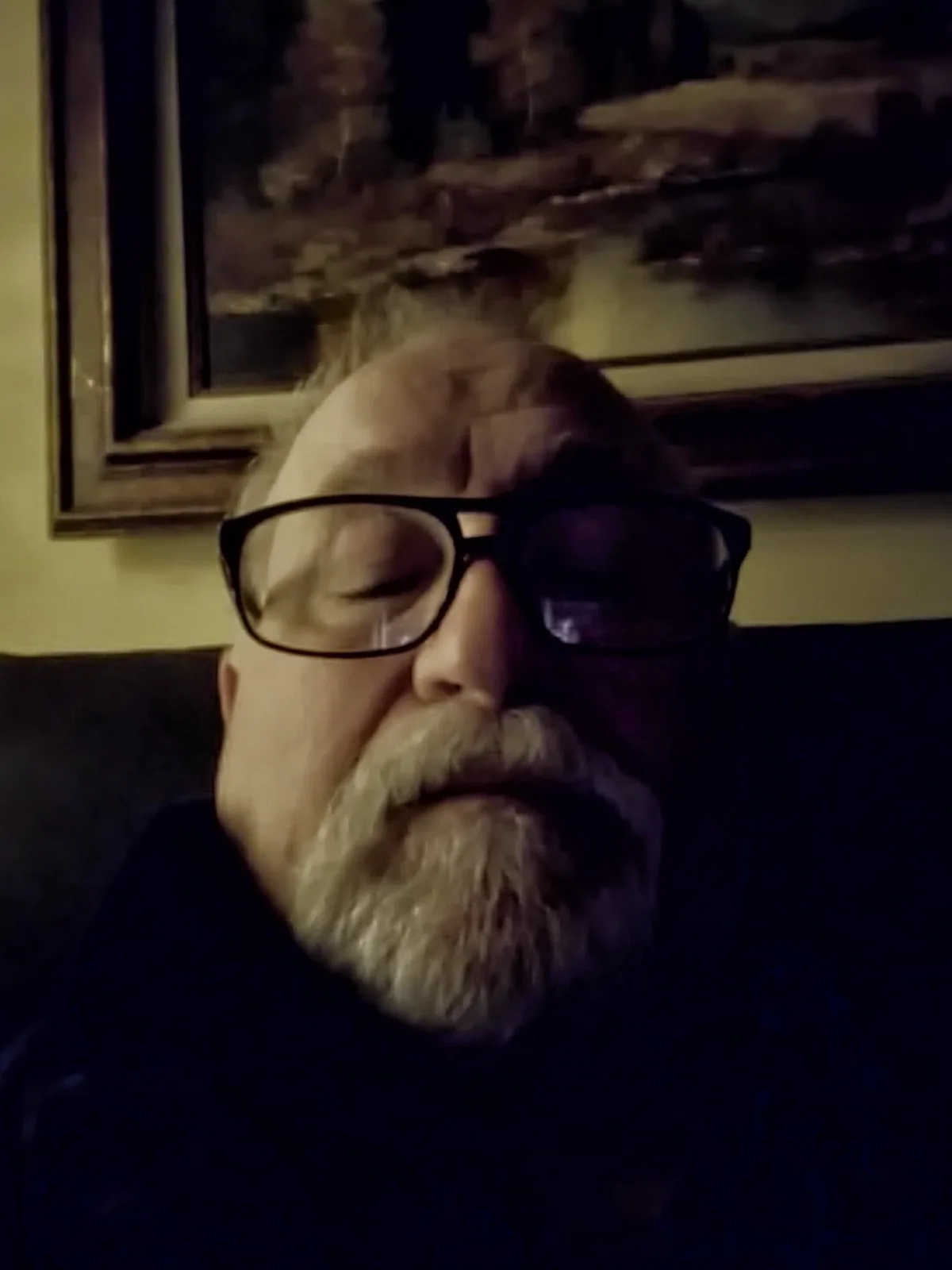A close-up photo of a middle-aged man with a white beard and glasses, eyes closed, appearing to be resting or asleep while…