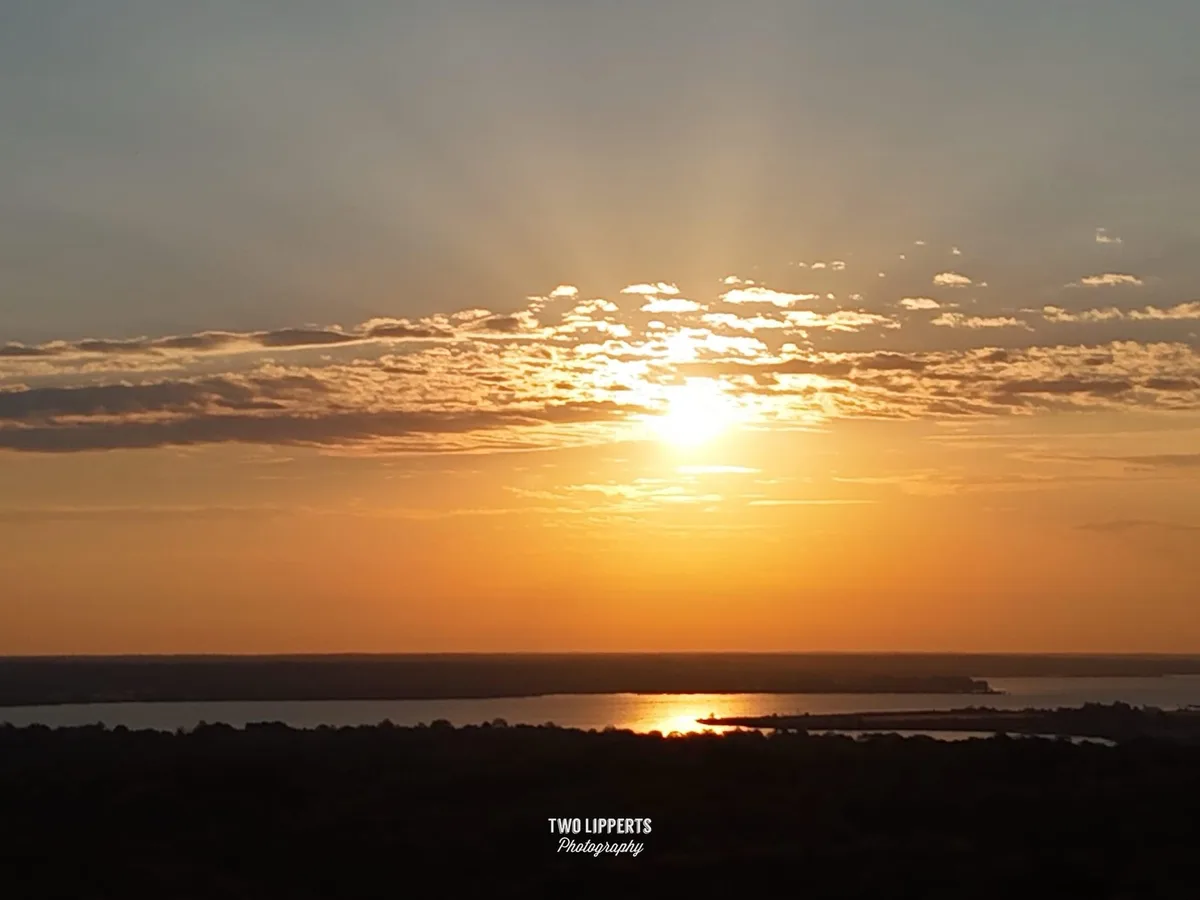 A serene sunset photograph showing the sun low on the horizon, casting golden light across scattered clouds and reflecting…