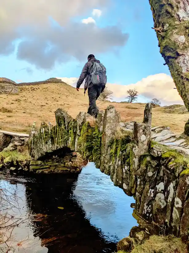 A person wearing a backpack walks across a narrow, moss-covered stone bridge over a stream in a rural landscape. The sky is…