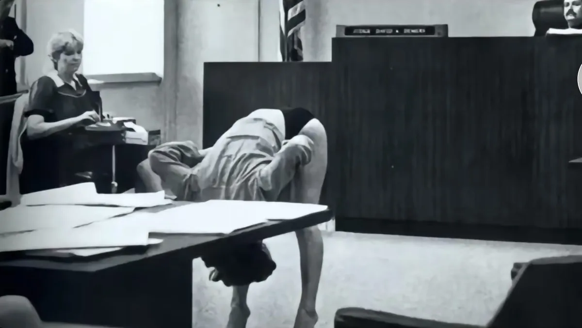 A black-and-white photo shows a person in a courtroom bending over a table with their head down, appearing to cry or be…