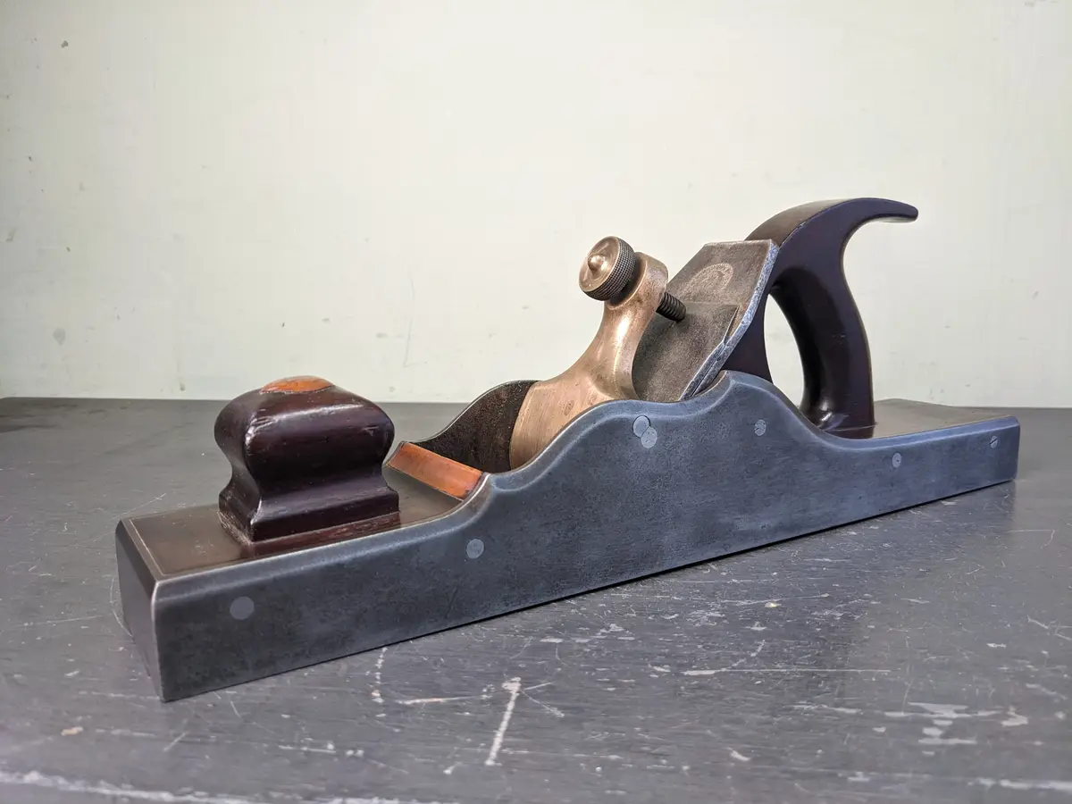 A vintage woodworking hand plane with a dark metal body, brass adjustment knob, and dark wooden handle and knob rests on a…