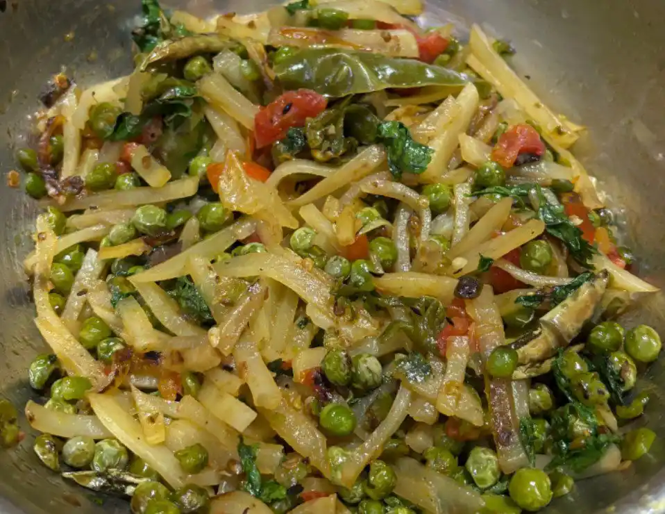 a close-up photo of a cooked vegetable dish in a metal bowl, featuring sliced potatoes, green peas, chopped tomatoes, and…