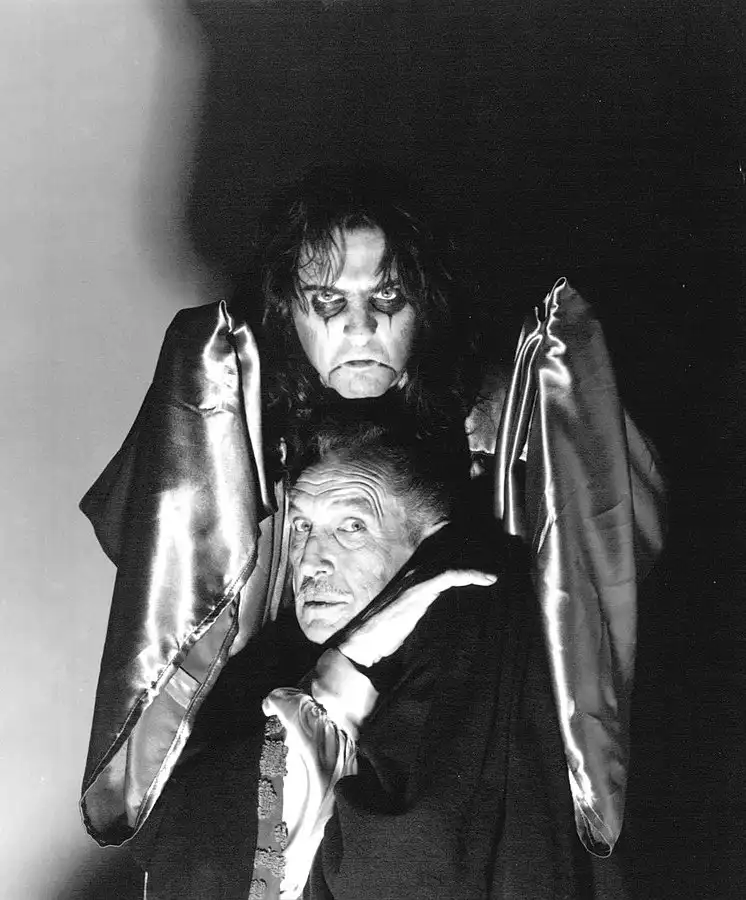 A black-and-white photo shows two men in theatrical vampire costumes. The man in the foreground has a startled expression…