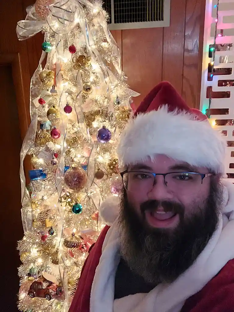 A man with a beard and glasses wearing a red and white Santa hat and a red Santa suit stands in front of a decorated, lit-up…