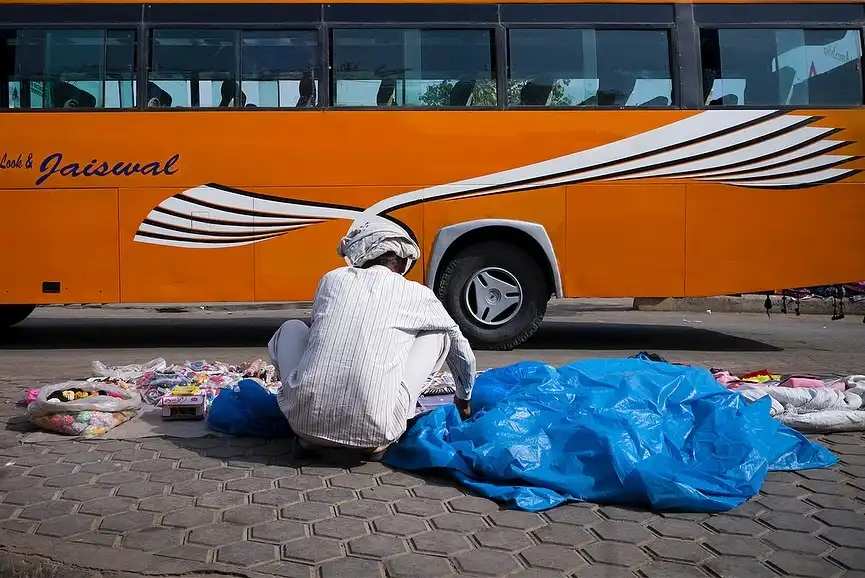 A person in a white striped shirt and head covering sits on a paved street, arranging colorful trinkets and toys on a blue…