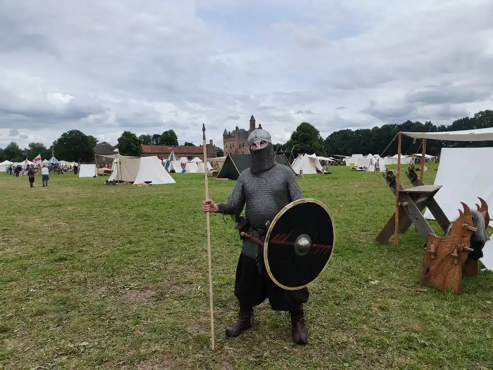 A person in full medieval chainmail armor, helmet, and face mask stands in a grassy field at what appears to be a historical…