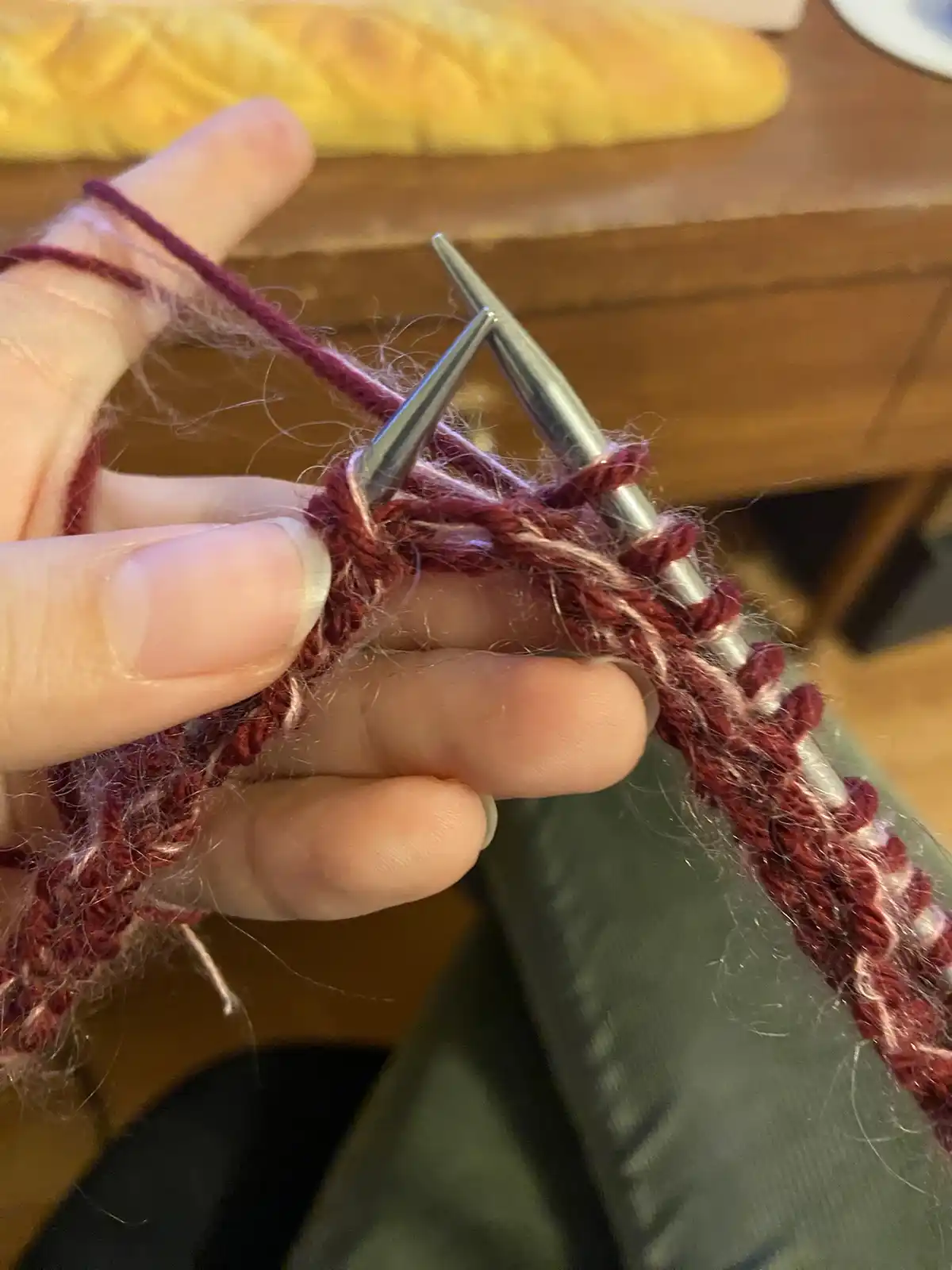 A close-up photo shows a person's hand holding two metal knitting needles with maroon and white yarn, actively knitting a…