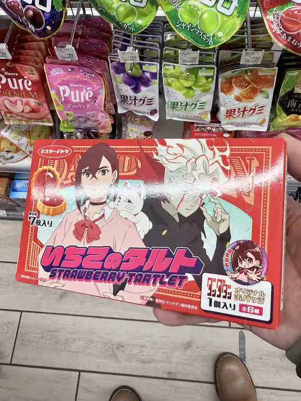 A hand holds a red box of 'Strawberry Tartlet' snacks featuring anime characters from 'Dandadan' in a Japanese supermarket…