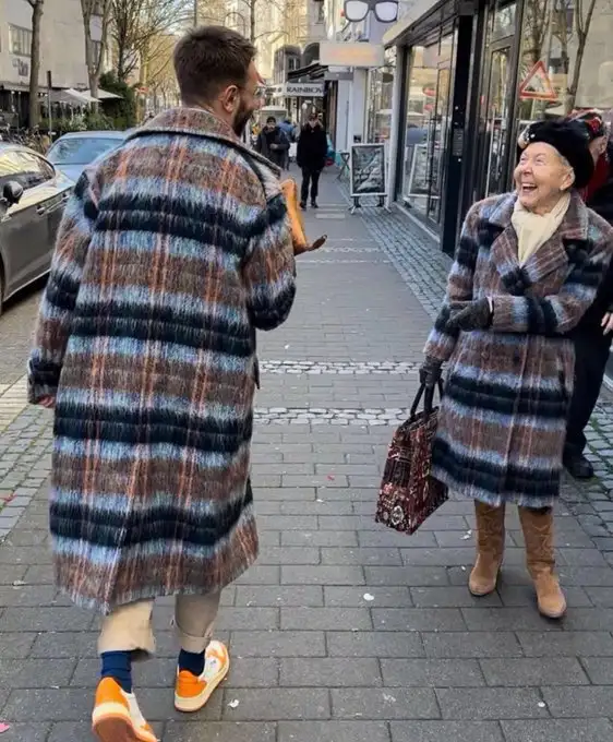 A man in a long, fuzzy, multicolored plaid coat walks away from an older woman who is laughing. The woman also wears a…